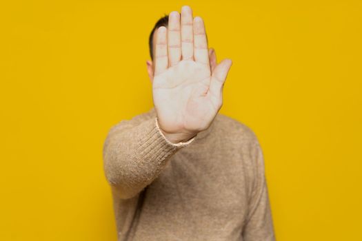 Hispanic man putting hand in front to stop camera, rejecting photos or images against yellow colored wall