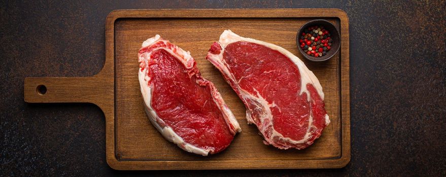 Two raw uncooked meat beef rib eye marbled steaks on wooden cutting board with seasonings on dark rustic background ready to be grilled from above, preparing dinner with meat