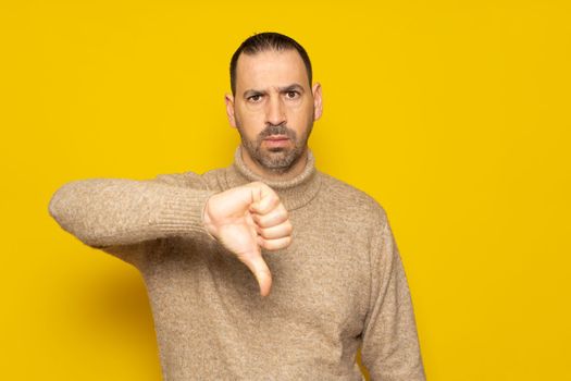 Hispanic man with a beard wearing a beige turtleneck posing with a serious expression over a yellow background has his thumbs down in disapproval