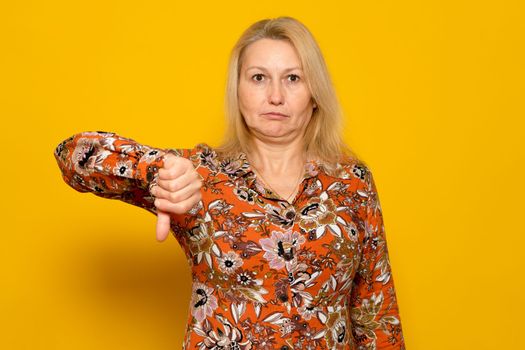 Blonde pretty woman wearing patterned dress over yellow background looking unhappy and angry showing rejection and negative thumbs down gesture. Bad expression.