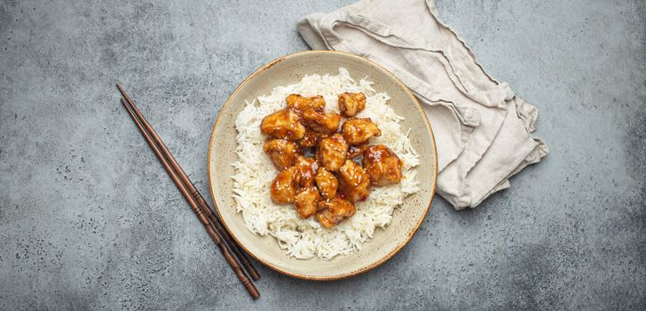 Chinese sweet and sour sticky chicken with sesame seeds and rice on ceramic plate with chopsticks top view on gray rustic stone background, traditional dish of China