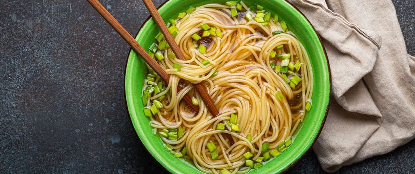 Asian noodles soup in green rustic ceramic bowl with wooden chopsticks top view on rustic stone background. Lo mein noodles with bouillon and green onion