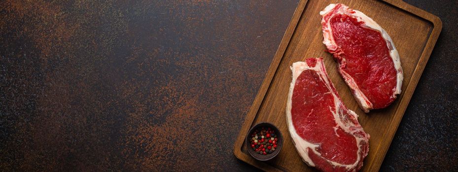 Two raw uncooked meat beef rib eye marbled steaks on wooden cutting board with seasonings on dark rustic background ready to be grilled from above, preparing dinner with meat, space for text