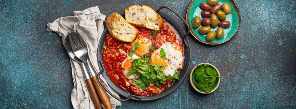 Middle Eastern and Maghrebi healthy dish Shakshouka made of eggs and tomato sauce served in pan with toasts, fresh cilantro and olives on rustic concrete background table from above