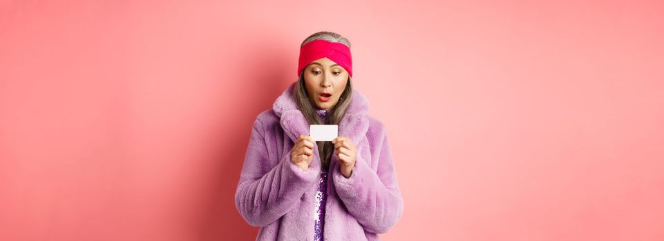 Shopping and fashion concept. Stylish asian mature woman looking at plastic credit card with amazement, standing in purple faux fur coat over pink background.