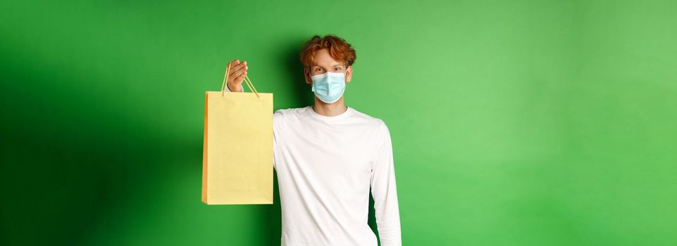 Portrait of handsome boyfriend in face mask giving shopping bag with valentines gift, smiling with eyes, standing over green background.