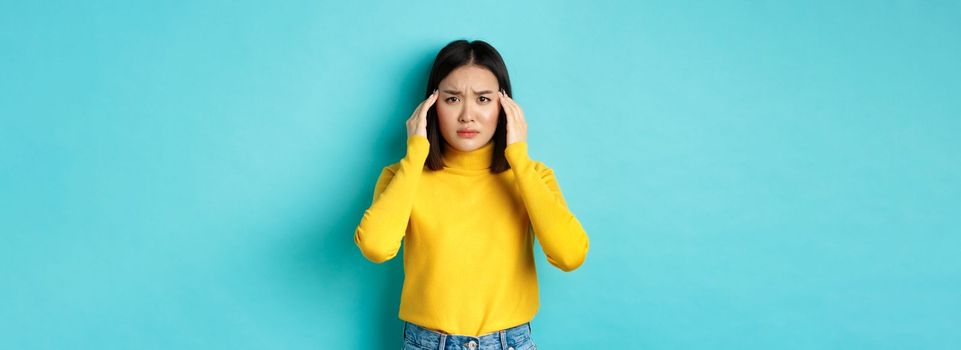 Image of distressed asian woman touching head and frowning, feeling headache, standing troubled against blue background.