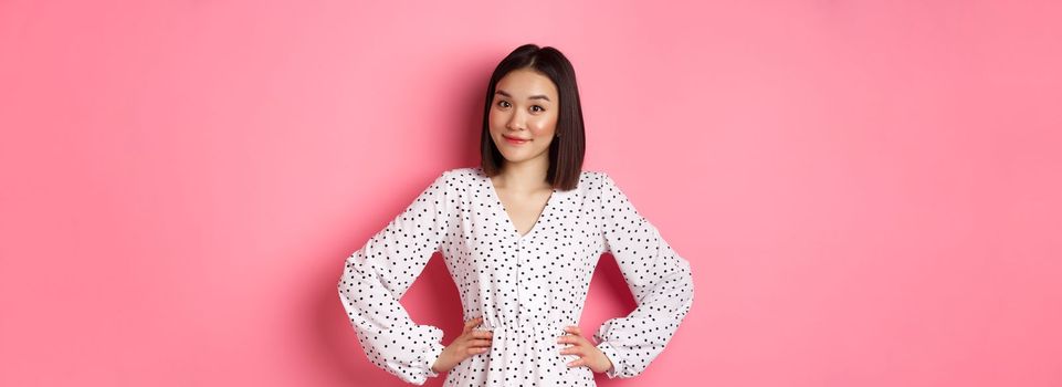 Beautiful korean lady in dress looking at camera and smiling, waiting for something, standing against pink background.