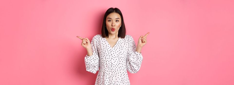 Beautiful asian woman making choice on shopping, pointing fingers sideways and showing variants, smiling at camera, standing over pink background.