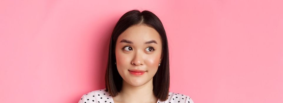 Beauty and skin care concept. Close-up of cute asian teenage girl looking left at banner, smiling silly, standing in dress over pink background.