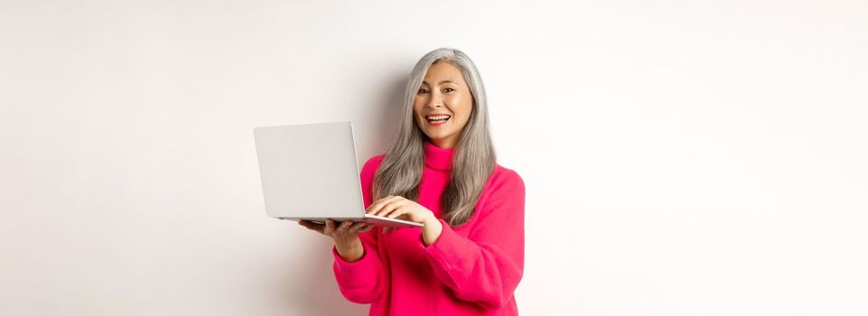 Beautiful asian senior woman entrepreneur working with laptop, laughing and smiling at camera, standing over white background.