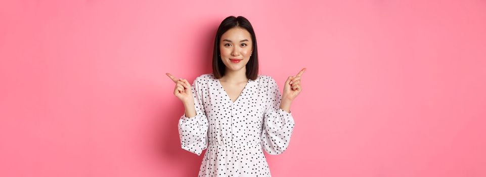 Beautiful asian woman making choice on shopping, pointing fingers sideways and showing variants, smiling at camera, standing over pink background.