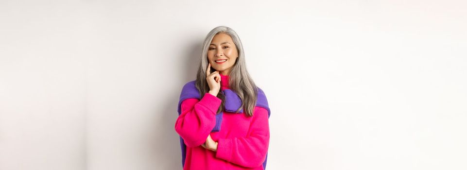 Fashionable asian middle-aged woman with gray hair, smiling pleased and thoughtful, having an idea, standing over white background.