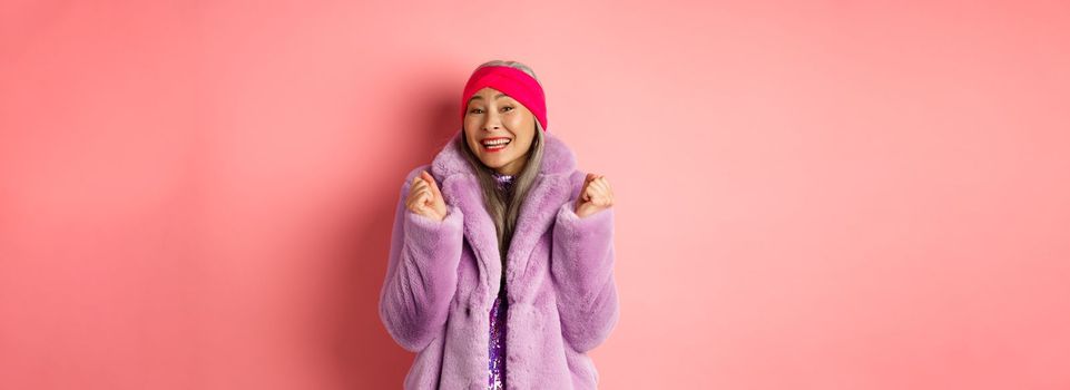 Cheerful asian lady in stylish funky coat, celebrating victory or success, saying yes and smiling happy, standing over pink background.