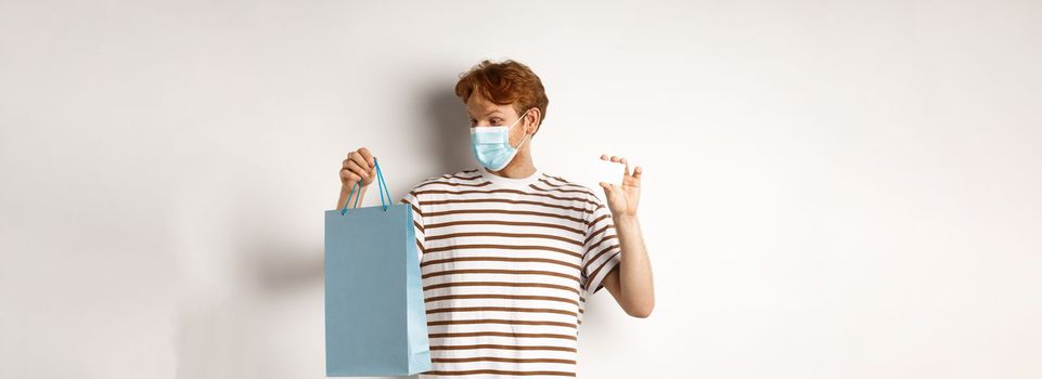 Concept of covid-19 and lifestyle. Cheerful young man with red hair, wear medical mask, showing shopping bag from store and plastic credit card.