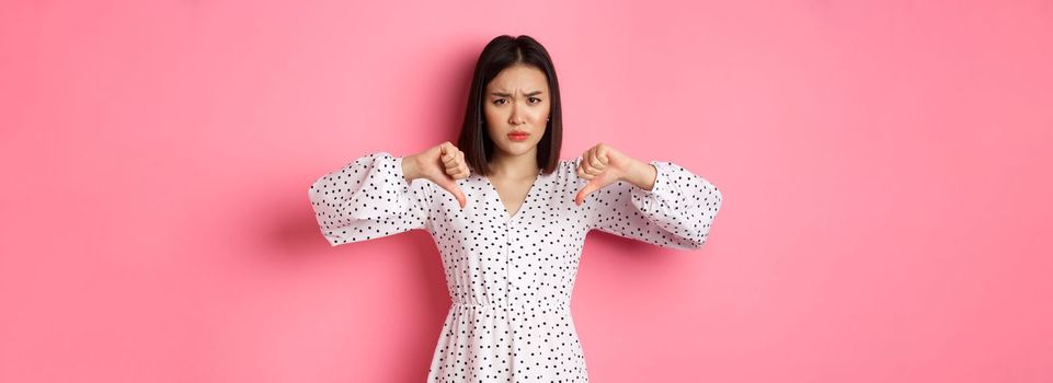 Disappointed asian woman showing thumbs-down, disapprove and dislike something, showing negative judgement, standing over pink background.