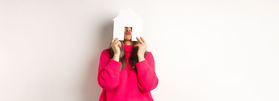 Real estate. Funny asian middle-aged woman hiding face behind paper house model and showing puckered lips, having fun, standing over white background.