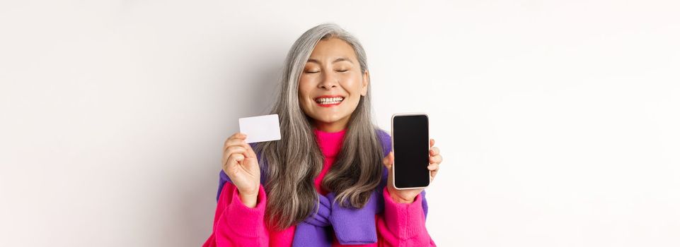 Online shopping. Happy and delighted asian senior woman smiling with joy, showing blank smartphone screen and plastic card, buying in internet, white background.