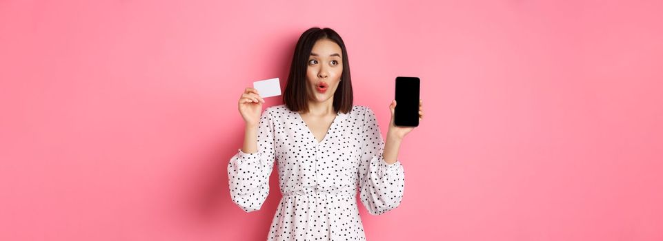 Cute asian woman shopping online, showing bank credit card and mobile screen, smiling and looking left at copy space, standing over pink background.