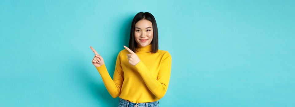 Shopping concept. Attractive young asian woman with perfect skin, wearing trendy yellow sweater and jeans, pointing fingers right and smiling, showing advertisement.