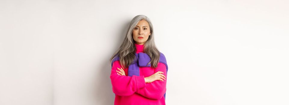 Serious and strict asian grandmother looking angry at camera, cross arms on chest, standing over white background disappointed.