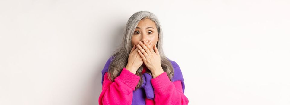 Close up of shocked senior lady gasping and covering mouth, staring at camera speechless, witness something, standing over white background.