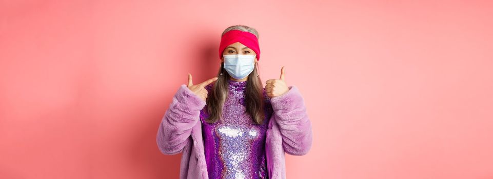 Covid-19, social distancing and fashion concept. Smiling asian senior woman in fashionable clothes and respirator, pointing at face mask and show thumb-up, pink background.