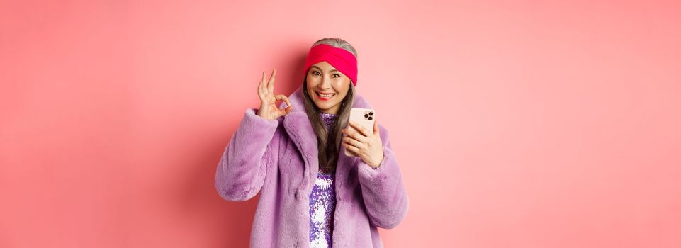 Online shopping and fashion concept. Stylish asian senior woman showing okay sign and holding mobile phone, recommending internet store, pink background.