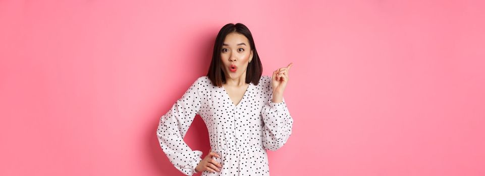 Excited asian female model showing promo offer, pointing at upper left corner and staring at camera amazed, wearing trendy spring dress, standing over pink background.