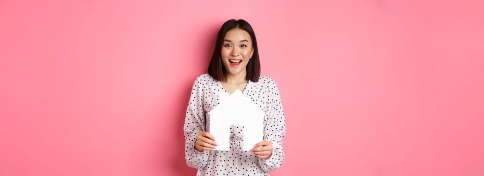 Real estate. Amazed asian woman searching for flat, holding house model and looking at camera happy, standing over pink background.