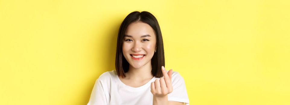 Valentines day and women concept. Close up of pretty asian girl in white t-shirt, smiling and showing finger heart, standing over yellow background.