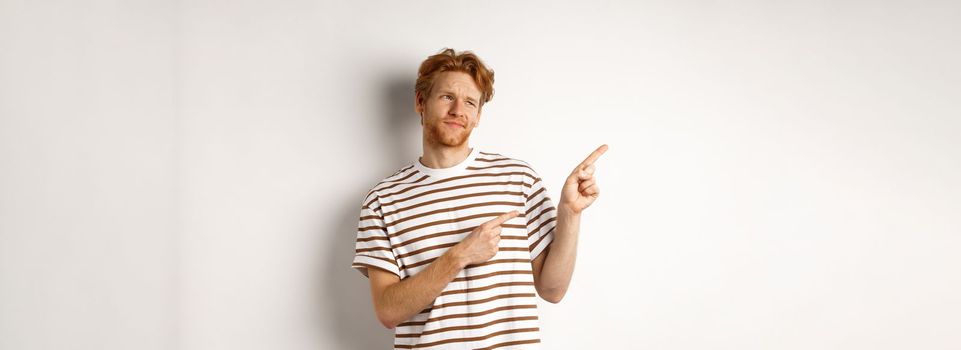 Doubtful young man with bristle and red hair looking, pointing fingers left at something average, staring skeptical at promo, white background.