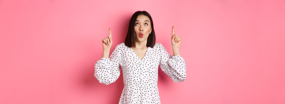 Cute korean girl in beautiful dress saying wow, looking and pointing fingers up, intrigued in promo offer, standing over pink background.