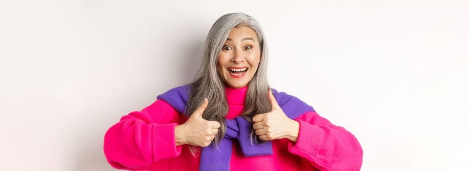 Excited and amazed asian elderly woman showing thumbs-up in approval, smiling happy at camera, praising something awesome, white background.