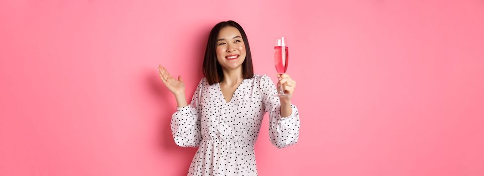 Beautiful asian woman, raising glass of champagne and smiling happy, drinking wine and celebrating, standing over pink background.