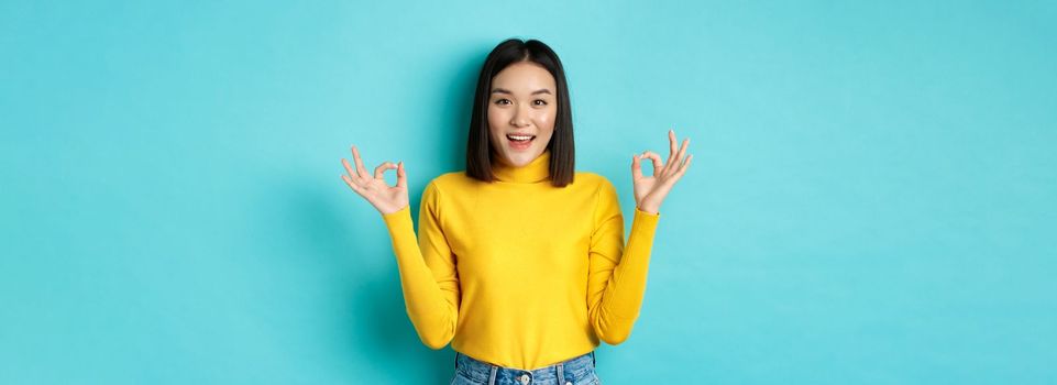 Cheerful asian female model showing okay gestures, smiling and looking impressed, praise product, standing over blue background.