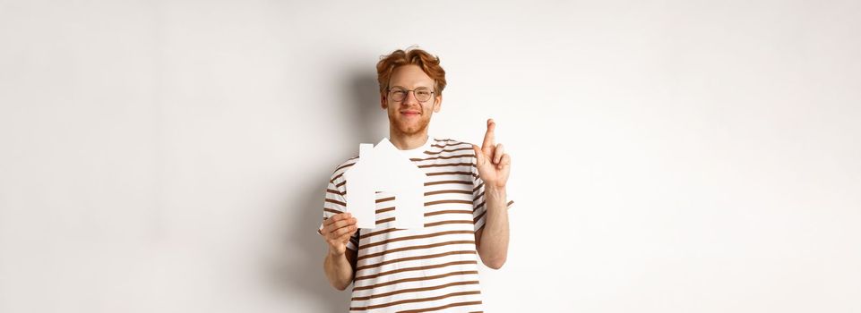Hopeful redhead man dreaming of buying house, holding paper home cutout and cross fingers for good luck, making a wish, standing over white background.