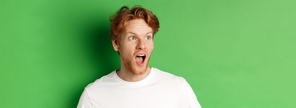 Close up of surprised and impressed redhead man checking out promotion offer, looking left with dropped jaw, standing over green background.