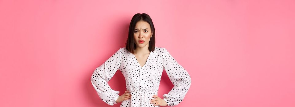 Angry asian woman staring at camera frustrated, holding hands on waist and waiting for explanations, standing in dress against pink background.