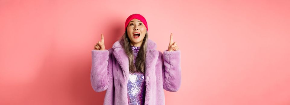 Mature asian hipster woman pointing fingers up, looking at top promo with amazement, checking out special deal, standing over pink background.