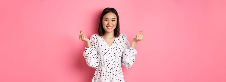 Lovely asian woman in dress showing korean heart signs and smiling, standing on romantic pink background.