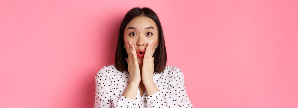 Close-up of surprised and amazed asian woman staring at camera, gasping impressed, holding hands near mouth, standing over pink background.