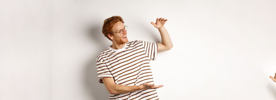 Happy and satisfied caucasian redhead man introduce something big, showing large product and smiling pleased, white background.