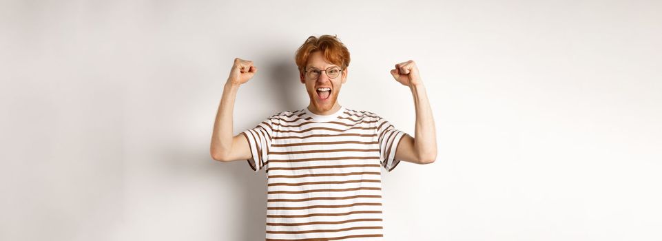 Joyful redhead guy raising hands up like winner, winning prize and celebrating, shouting for joy, standing over white background.