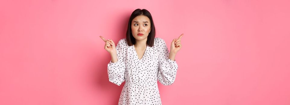 Confused asian woman hesitating, shrugging and pointing hands two sides, staring indecisive and need help with choice, standing over pink background.
