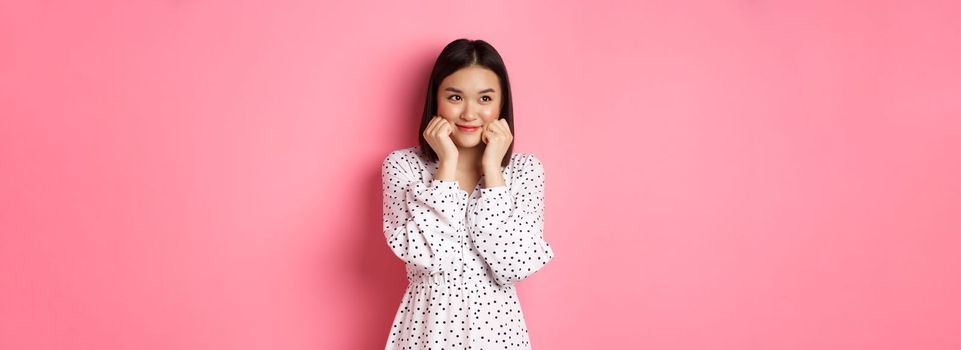 Cute and shy asian girl blushing, touching cheeks and looking left at copy space silly, standing against pink background.