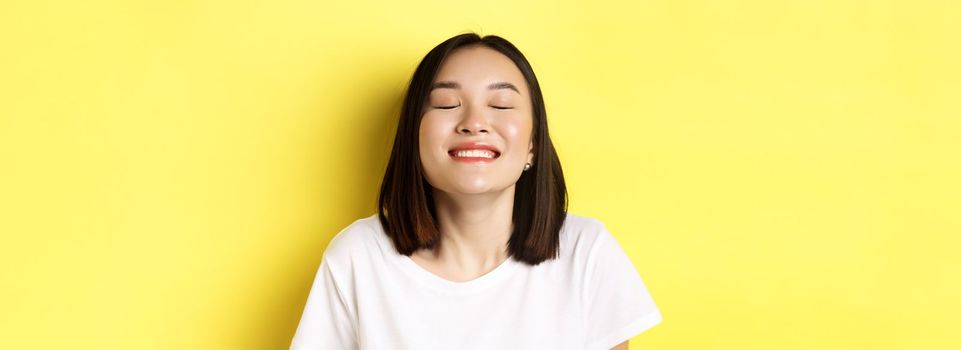 Close up of happy and relaxed asian woman enjoying sun, smiling with eyes closed and looking joyful, standing over yellow background.