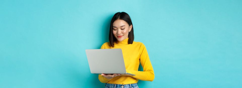 Beautiful asian female student working on laptop, typing on keyboard and looking at screen with pleased smile, standing over blue background.