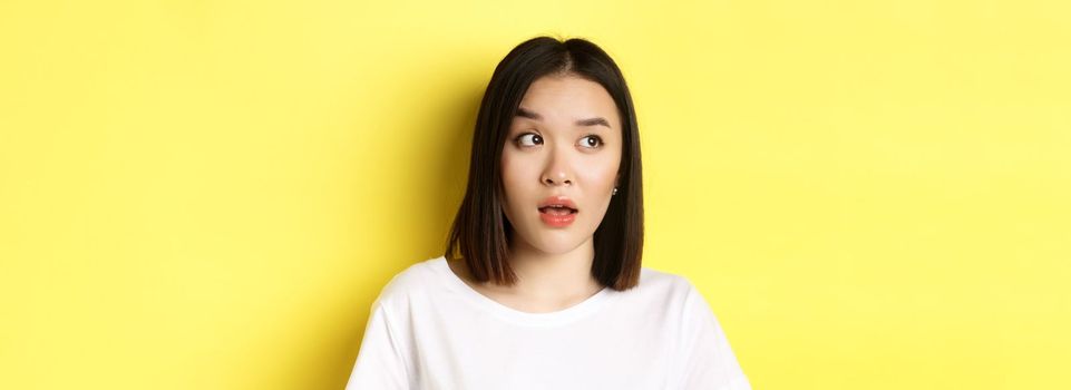 Close up of beautiful asian woman with clean perfect skin, raising eyebrow and looking left with intrigued face, standing over yellow background.