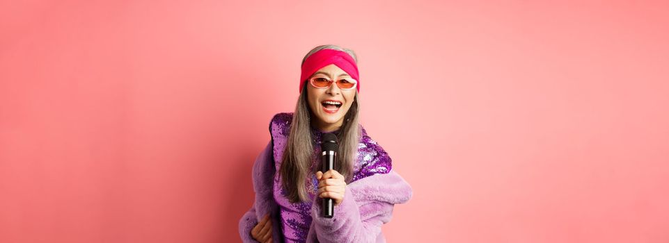 Fashion and lifestyle concept. Beautiful middle-aged woman in sunglasses, party dress and faux fur coat, singing in microphone and having fun at karaoke bar, pink background.